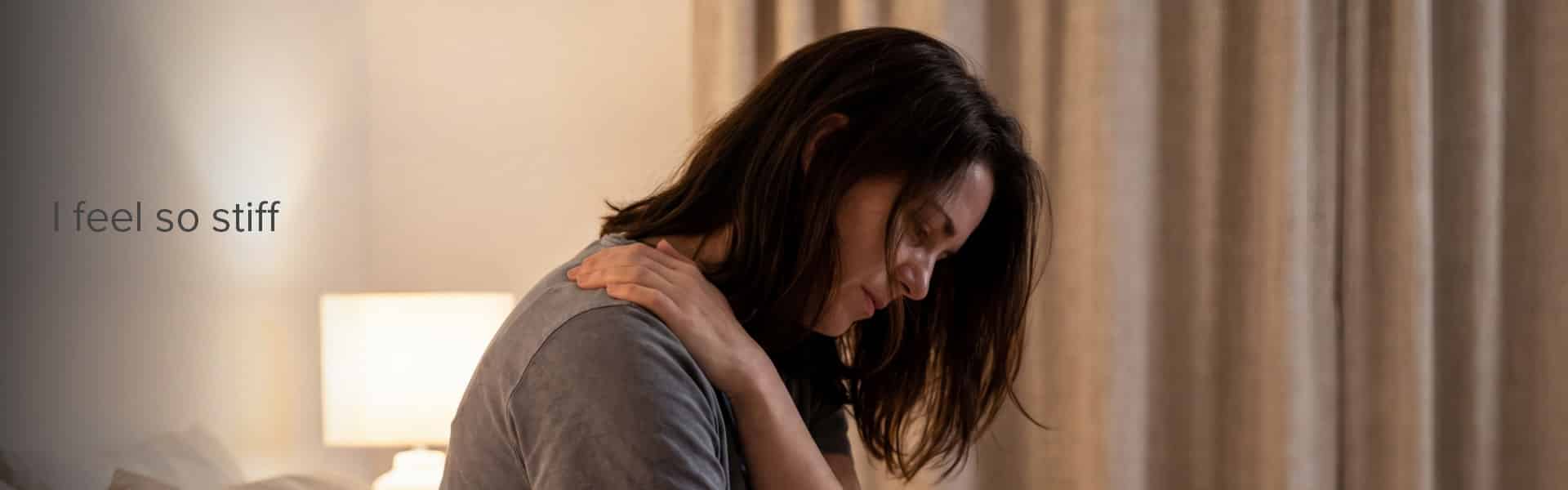 Woman sitting on bed holding shoulder, appearing stiff or uncomfortable