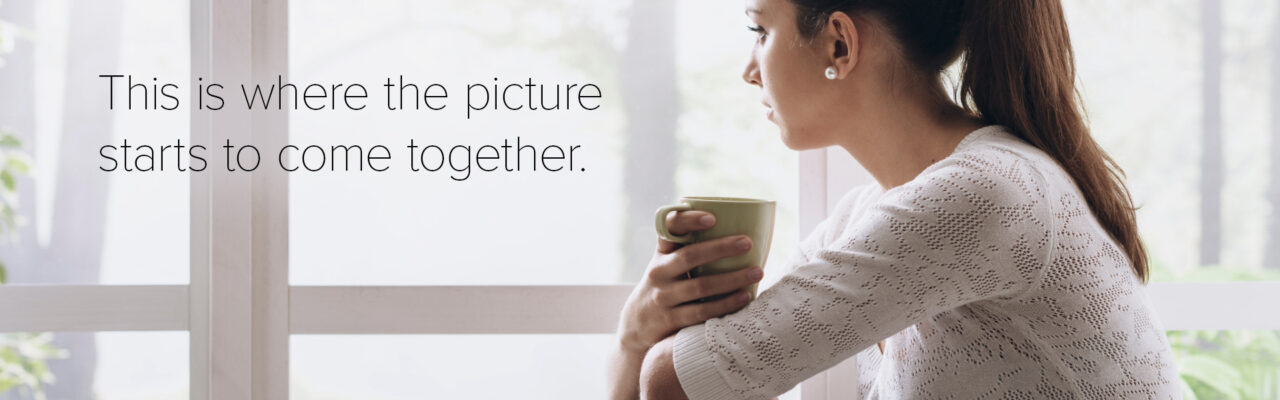 A woman sitting by a window holding a mug, looking to the side with a neutral expression.